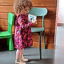 child, toddler, curly_hair, dress, pink_tights, boots, book, reading, indoor, wooden_wall, green_chair, blue_chair, floor, standing, person, colorful_clothing, young_child, playroom, expression, furniture