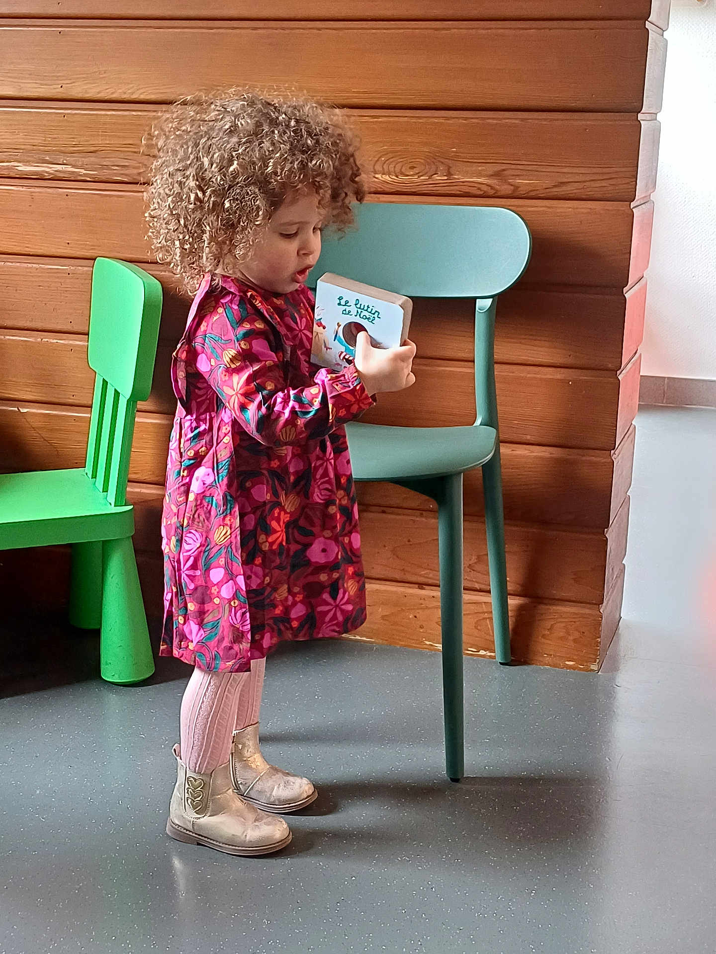 Lina participe au concours pour gagner de l'argent avec cette photo : child, toddler, curly_hair, dress, pink_tights, boots, book, reading, indoor, wooden_wall, green_chair, blue_chair, floor, standing, person, colorful_clothing, young_child, playroom, expression, furniture