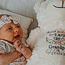 baby, infant, child, portrait, lying_down, smiling, happy, floral_outfit, headband, teddy_bear, stuffed_animal, bed, pillow, blanket, nursery, indoor, cozy, embroidered_text, striped_background, closeup