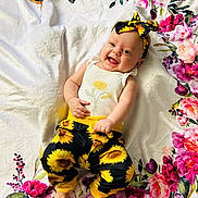 Joyce is registered to the contest to win money with this photo: baby, child, smiling, flower_pattern, headband, sunflower, pants, white_shirt, blanket, floral, happy, cute, infant, playful, indoors, colorful, person, portrait, lying_down, cheerful