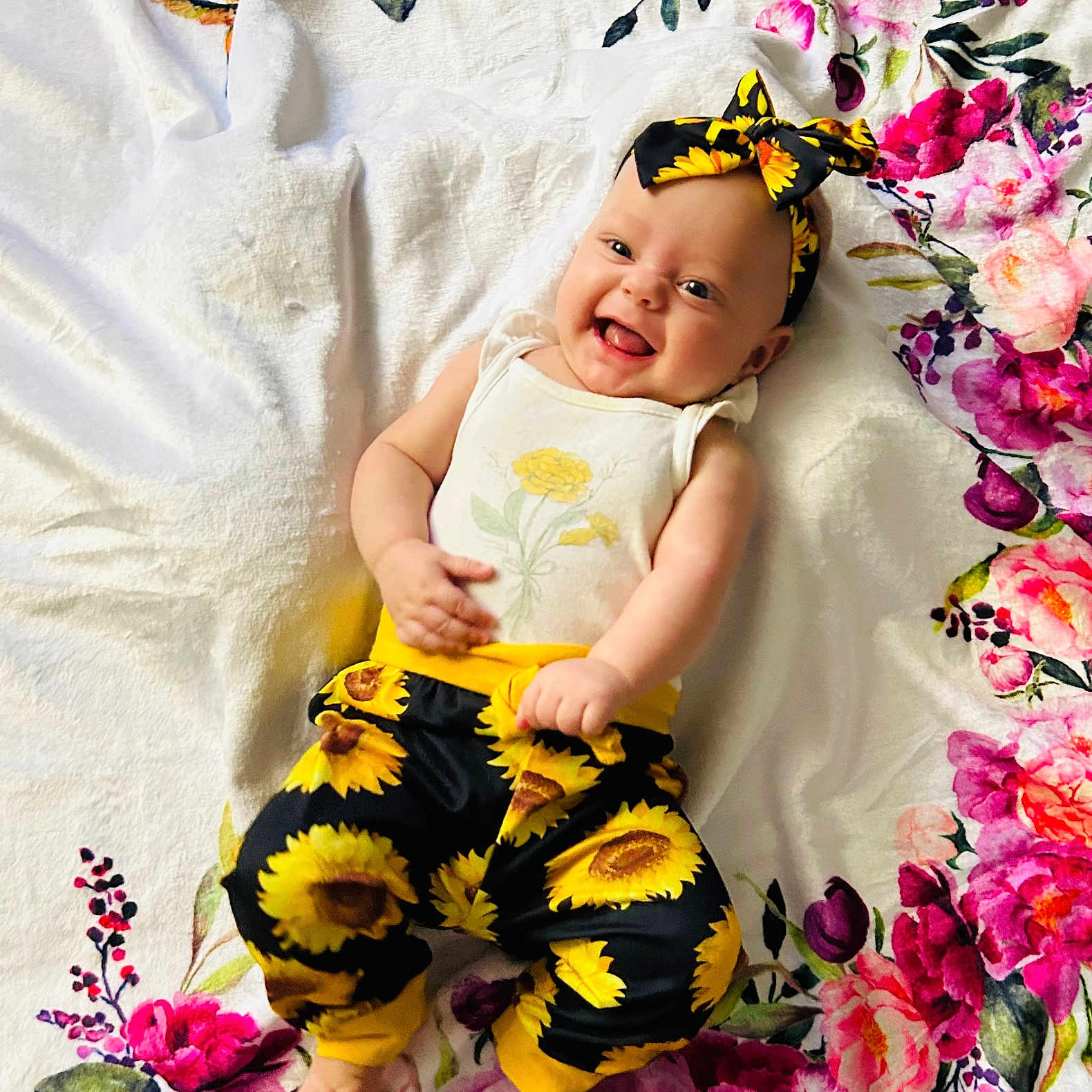 Joyce is registered to the contest to win money with this photo: baby, blanket, cheerful, child, colorful, cute, floral, flower_pattern, happy, headband, indoors, infant, lying_down, pants, person, playful, portrait, smiling, sunflower, white_shirt