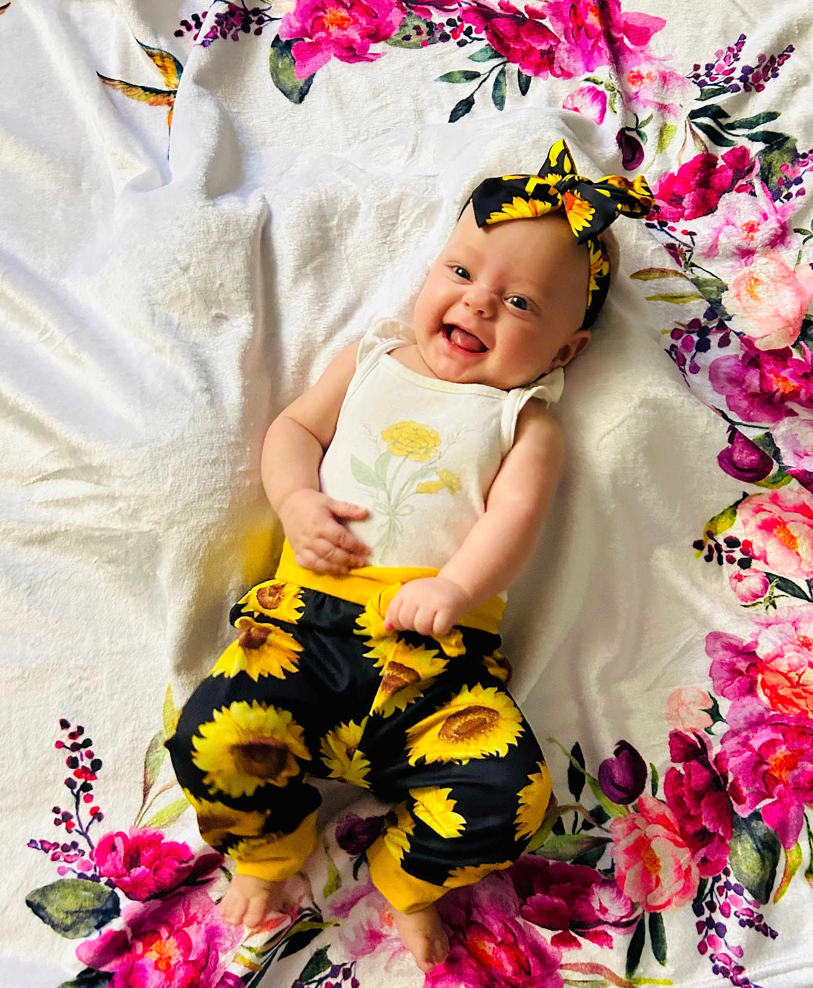 Joyce is registered to the contest to win money with this photo: baby, child, smiling, flower_pattern, headband, sunflower, pants, white_shirt, blanket, floral, happy, cute, infant, playful, indoors, colorful, person, portrait, lying_down, cheerful