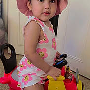 Maddie is registered to the contest to win money with this photo: toddler, child, pink_hat, floral_romper, toy_airplane, indoor, carpet, mickey_mouse, wheels, yellow, red, smiling, cute, play, hat, face, person, young_child, home, fun