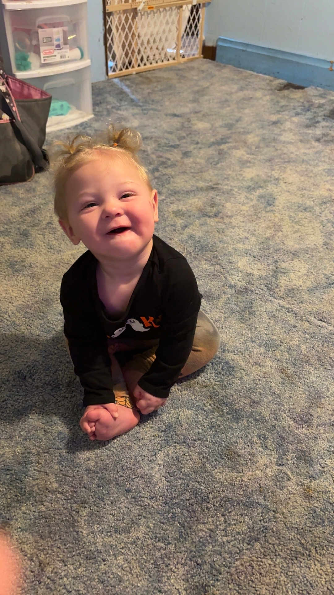 Isabella joined the competition — help win amazing prizes! toddler, child, smiling, sitting, carpet, indoor, baby_gate, storage_drawer, black_shirt, blond_hair, pigtails, happy, person, floor, room, casual_clothing, footwear, playful, cute, young_child