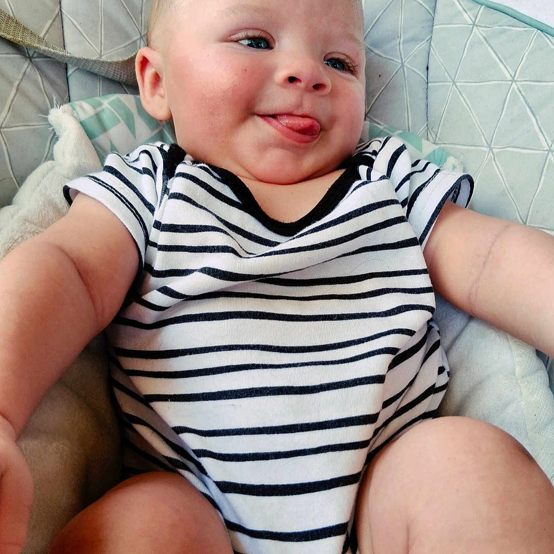 Caleb joined the competition — help win amazing prizes! baby, child, infant, striped_clothing, onesie, tongue_out, smiling, cushion, pillow, soft_furniture, indoor, closeup, happy, face, skin, arm, leg, head, seated, person