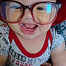 baby, child, glasses, red_hat, smiling, happy, hand, clothing, onesie, indoors, portrait, cute, person, face, headwear, accessory, skin, fingers, joy, human