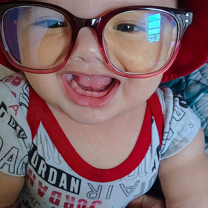 Jesse is registered to the contest to win money with this photo: baby, child, glasses, red_hat, smiling, happy, hand, clothing, onesie, indoors, portrait, cute, person, face, headwear, accessory, skin, fingers, joy, human