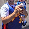 baby, basketball_jersey, blue_hat, casual_clothing, child, daytime, dog, family, greenery, happy, holding, man, outdoor, person, portrait, smile, street, sunlight, urban, wall