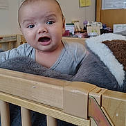 Jesse is registered to the contest to win money with this photo: baby, crib, stuffed_animal, wooden_crib, indoor, child, expression, curious, soft_toy, furniture, room, home, infant, face, person, cute, cozy, portrait, young_child