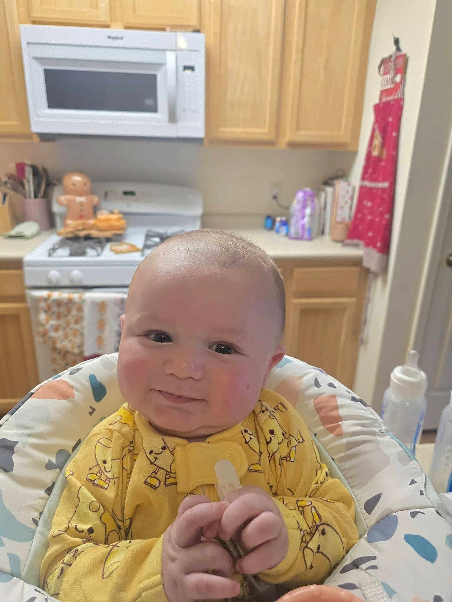 Charlie joined the competition — help win amazing prizes! baby, infant, smile, yellow_onesie, baby_seat, kitchen, stove, gingerbread_man, cabinet, microwave, countertop, baby_bottle, happy, cute, indoor, child, person, face, hand, home