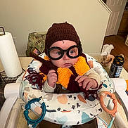 Charlie is registered to the contest to win money with this photo: baby, child, glasses, hat, scarf, knitted, wizard, costume, baby_seat, toy, indoor, person, face, curious, clothing, brown_pants, table, kitchen, paper_towel, can