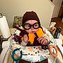 baby, child, glasses, hat, scarf, knitted, wizard, costume, baby_seat, toy, indoor, person, face, curious, clothing, brown_pants, table, kitchen, paper_towel, can