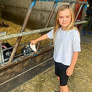 Daisy joined the competition — help win amazing prizes! child, girl, blonde_hair, white_shirt, black_shorts, baby_goat, goat, petting, barn, straw, fence, animal, farm, outdoor, smile, footwear, casual_clothing, daylight, young, nature
