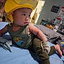 Celine is registered to the contest to win money with this photo: adult, baby, bed, blue_bed_sheet, cartoon, casual, child, curious, expression, face, furniture, helmet, indoor, pants, person, room, smiling, tank_top, toy, yellow
