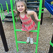 Scarlett is registered to the contest to win money with this photo: child, girl, playground, play_equipment, smiling, happy, outdoor, wood_chips, green_bars, dress, shoes, socks, daylight, fun, recreation, climbing, park, young_child, casual_clothing, joy