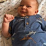 baby, infant, smile, blue_shirt, dinosaur_print, blanket, lying_down, cute, child, young, portrait, happy, indoors, soft_light, cozy, small_hand, skin, face, eyes, buttoned_shirt