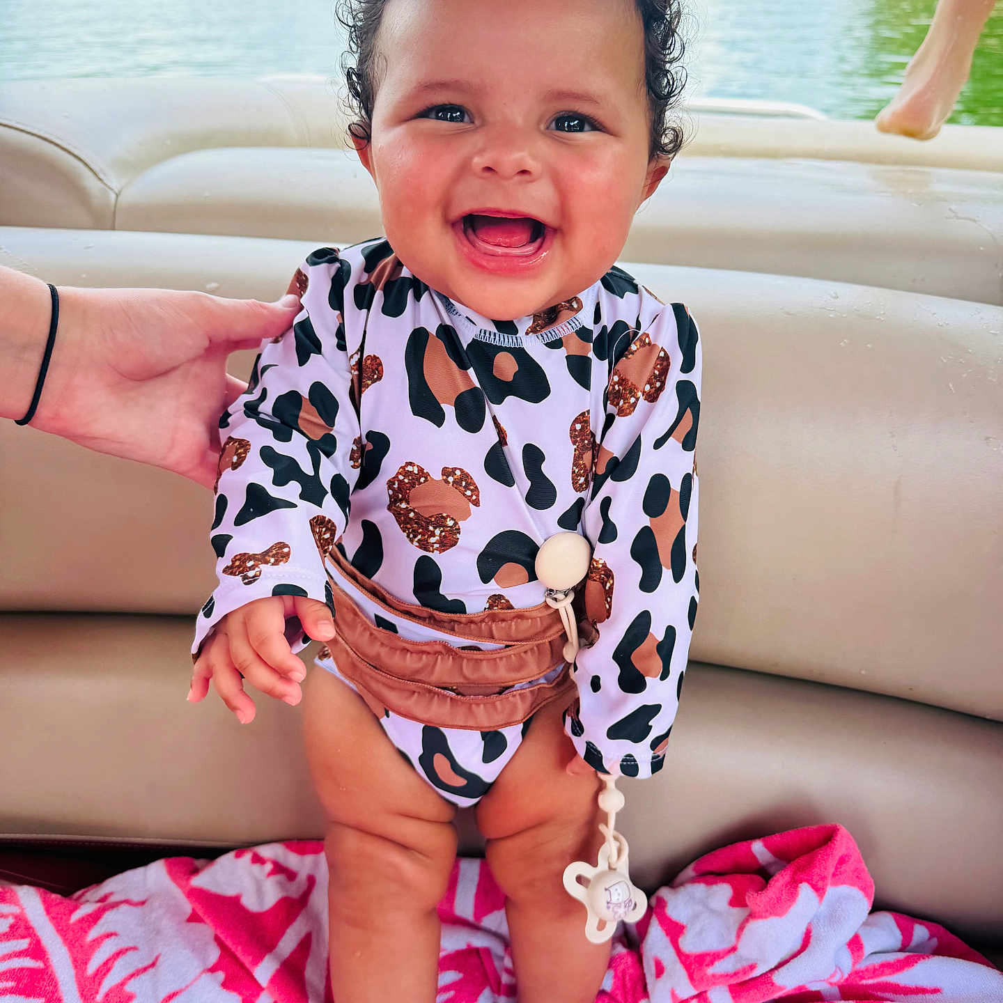 Lylah is registered to the contest to win money with this photo: baby, boat, child, curly_hair, greenery, hand, happy, infant, lake, leopard_print, nature, outdoor, person, pink, smiling, standing, summer, swimsuit, towel, water