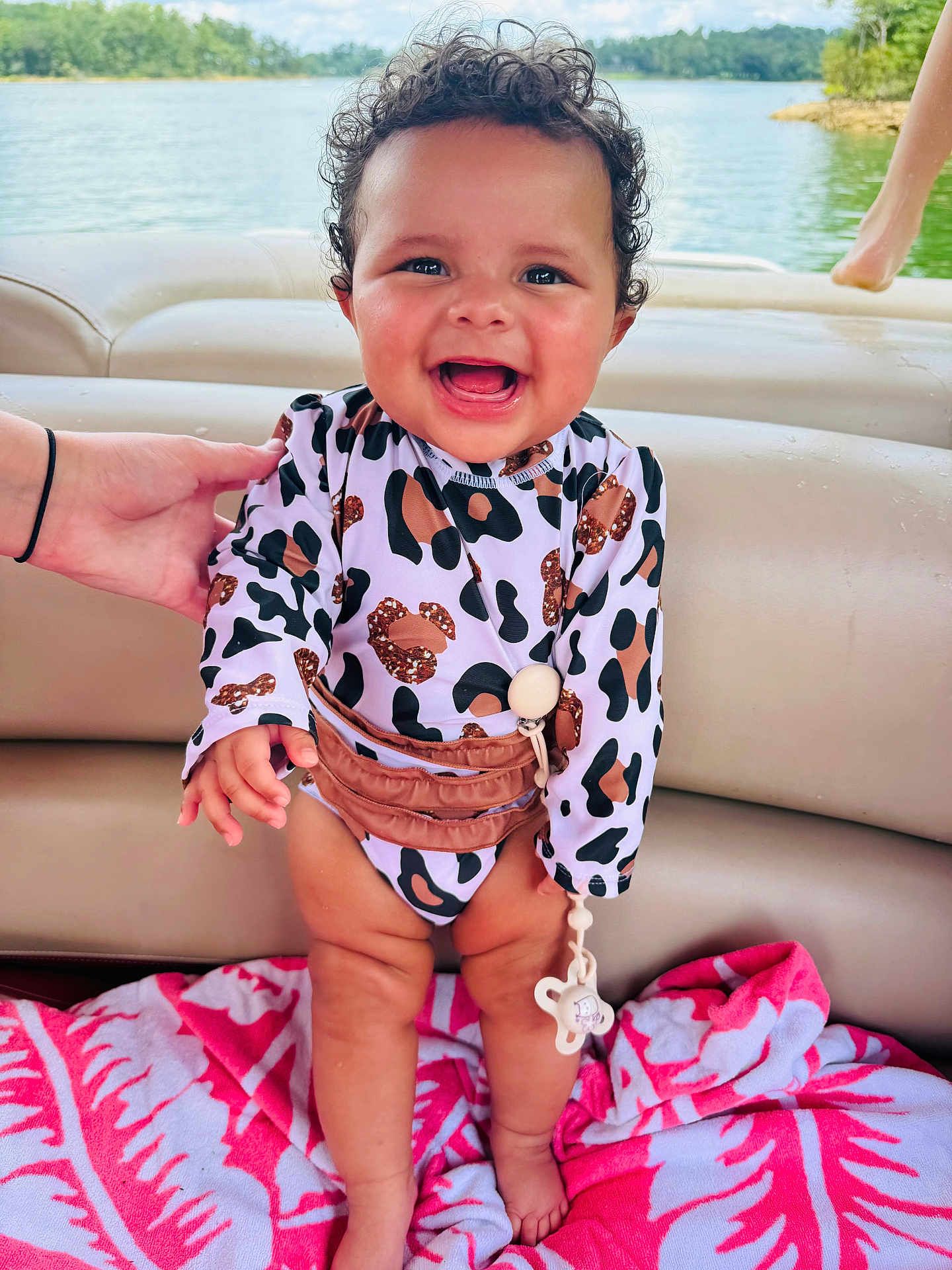 Lylah is registered to the contest to win money with this photo: baby, smiling, curly_hair, leopard_print, swimsuit, boat, lake, water, nature, greenery, hand, towel, pink, child, outdoor, happy, standing, person, infant, summer