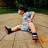 toddler, child, outdoor, greenery, stone_slab, curious, sitting, shorts, shirt, black_shoes, flame_pattern, grass, nature, playful, casual, young_child, park, daylight, side_view, bare_legs