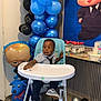 toddler, high_chair, balloons, party, blue, black, child, shirt, tie, shoes, floor, table, bottle, decoration, plastic, indoor, celebration, cartoon, toy, curious