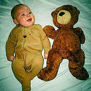 Albert is registered to the contest to win money with this photo: baby, teddy_bear, toy, bed, sleepwear, infant, smiling, cute, child, soft_toy, lying_down, white_sheet, brown_clothing, plush, comfort, indoors, person, portrait, happy, cozy