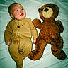 baby, teddy_bear, toy, bed, sleepwear, infant, smiling, cute, child, soft_toy, lying_down, white_sheet, brown_clothing, plush, comfort, indoors, person, portrait, happy, cozy