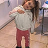 child, girl, indoor, tile_floor, jacket, pink_pants, adidas, long_hair, hair_tied, standing, pose, kitchen, cabinet, chair, footwear, curious, playful, head_tilt, person, floor