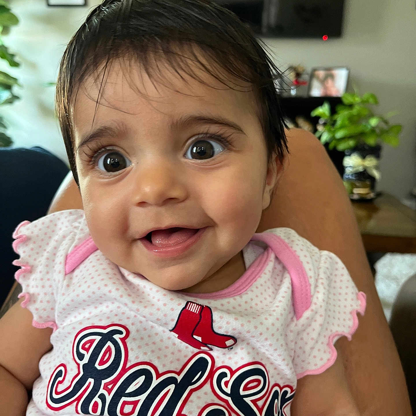 Priya is registered to the contest to win money with this photo: baby, child, clothing, cute, face, furniture, happy, home, human, indoor, infant, person, plant, portrait, red_sox, shirt, sitting, smiling, television, text