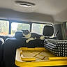 cat, black_and_white_cat, animal, pet, car, vehicle, back_seat, interior, storage_bin, yellow_lid, pet_bed, cushion, windows, sunlight, outdoor_view, lounging, sitting, whiskers, fur, road_trip
