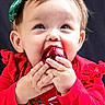 baby, child, face, hands, red_clothing, green_bow, christmas_ornament, holiday, festive, cute, portrait, smiling, indoors, holiday_season, santa_graphic, headband, closeup, cheerful, celebration, winter