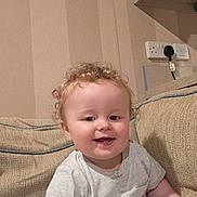 Noah is registered to the contest to win money with this photo: toddler, child, smiling, curly_hair, indoor, couch, beige, shirt, happy, face, person, plush, home_interior, wallpaper, power_outlet, light, seated, cute, skin, baby