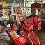 child, rocking_horse, toy, night, water_fountain, lights, holiday_decorations, cityscape, outdoor, play, smile, girl, clothing, boots, pavement, festive, building, window, raindrops, fun