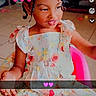 Daniya is registered to the contest to win money with this photo: bow, braids, child, cute, dress, floor, floral_pattern, girl, hair_accessory, heart_emoji, indoors, pink_chair, playful, portrait, seated, smiling, steering_wheel, text_overlay, toy, young_child