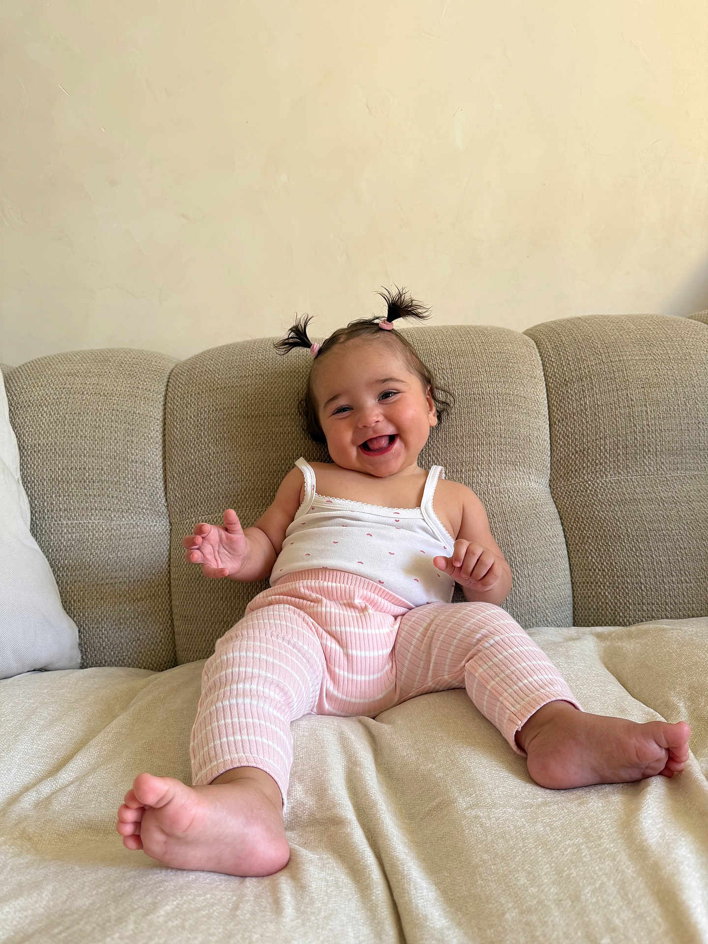 Isée participe au concours pour gagner de l'argent avec cette photo : baby, child, smiling, happy, pigtails, pink_pants, white_tank_top, couch, beige, indoor, person, cute, infant, small_hands, barefoot, plush, relaxed, portrait, sitting, home
