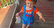 Rhiggs is registered to the contest to win money with this photo: child, toddler, barefoot, overalls, orange_shirt, cap, candy, wooden_porch, outdoor, person, hand, foot, bag, railing, window, reflection, floor, sunlight, casual, curious