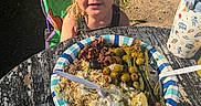blonde_hair, chair, cheese, child, food, fork, grass, green_beans, olives, outdoor, picnic, plastic_utensil, plate, potato_salad, rustic, shadow, smiling, sunlight, table, water_bottle