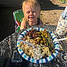 Rhiggs is registered to the contest to win money with this photo: child, blonde_hair, smiling, outdoor, food, plate, fork, table, green_beans, olives, cheese, potato_salad, water_bottle, sunlight, shadow, grass, chair, picnic, rustic, plastic_utensil