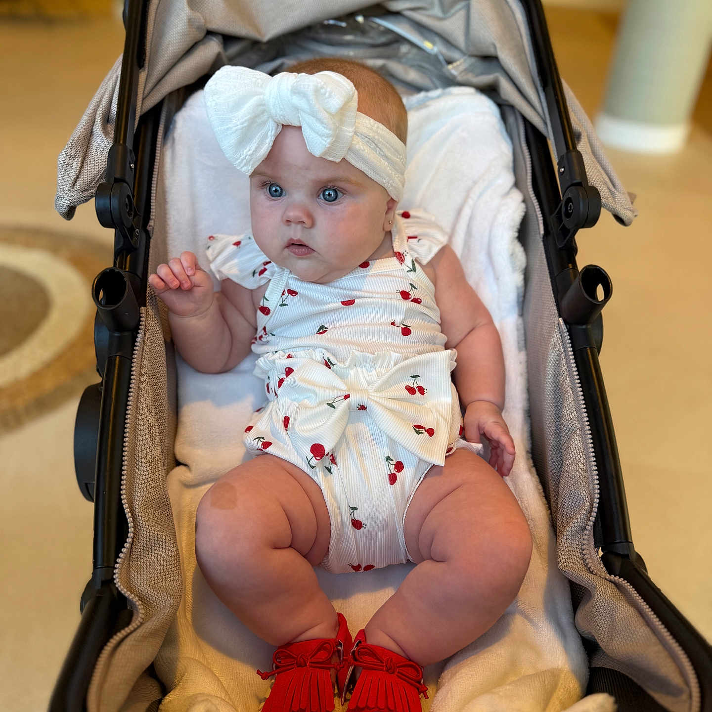 Billie J is registered to the contest to win money with this photo: baby, bow, carriage, cherry_pattern, child, clothing, curious, cute, footwear, headband, indoor, infant, person, plush_blanket, red_shoes, sitting, soft_lighting, stroller, white_outfit, young_child