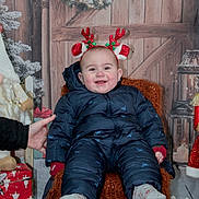 Maellan participe au concours pour gagner de l'argent avec cette photo : baby, child, smiling, winter_clothing, snowsuit, headband, antlers, holiday, christmas, decorations, wreath, chair, hand_support, cozy, indoor, festive, red, blue, wooden_background, smile