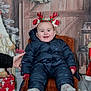 baby, child, smiling, winter_clothing, snowsuit, headband, antlers, holiday, christmas, decorations, wreath, chair, hand_support, cozy, indoor, festive, red, blue, wooden_background, smile
