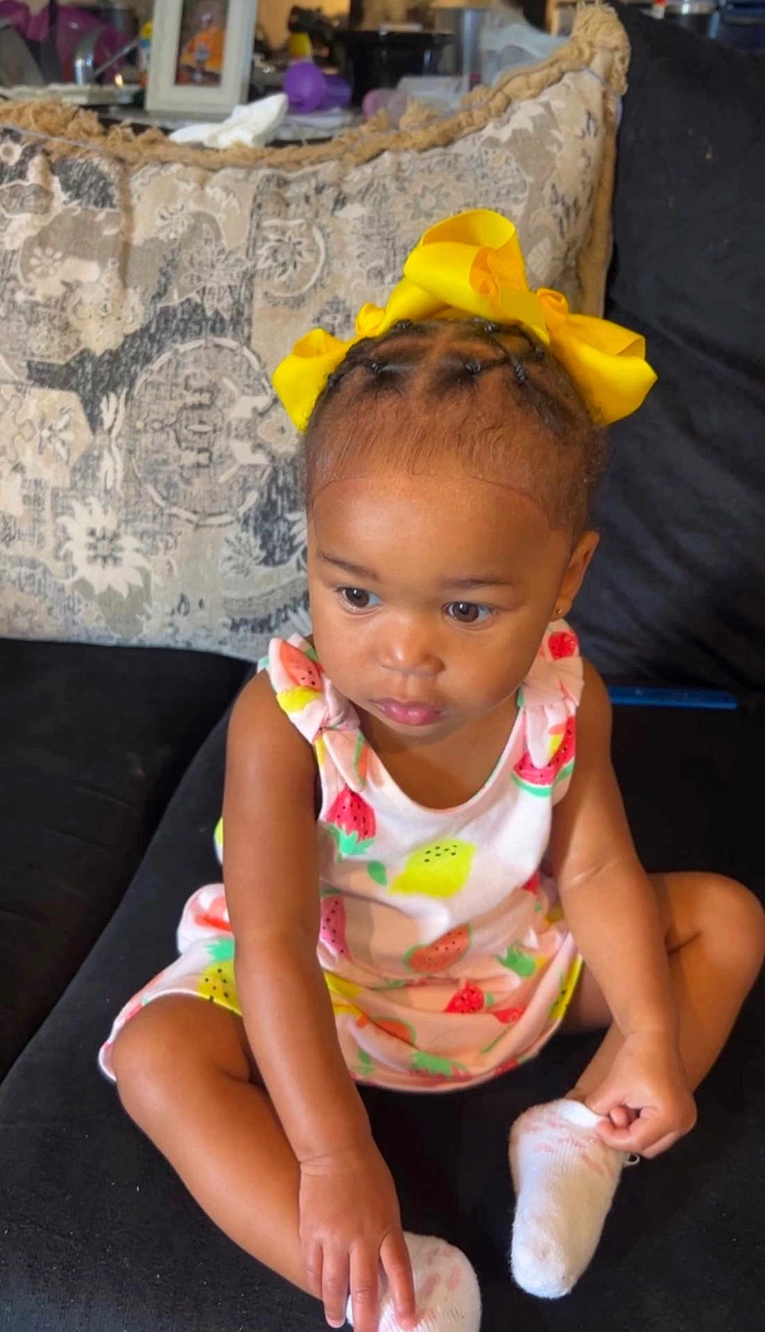 Alayla joined the competition — help win amazing prizes! toddler, child, girl, yellow_bow, braided_hair, dress, fruit_pattern, socks, couch, indoor, curious, sitting, person, young_child, black_couch, home, portrait, cute, fashion, expression