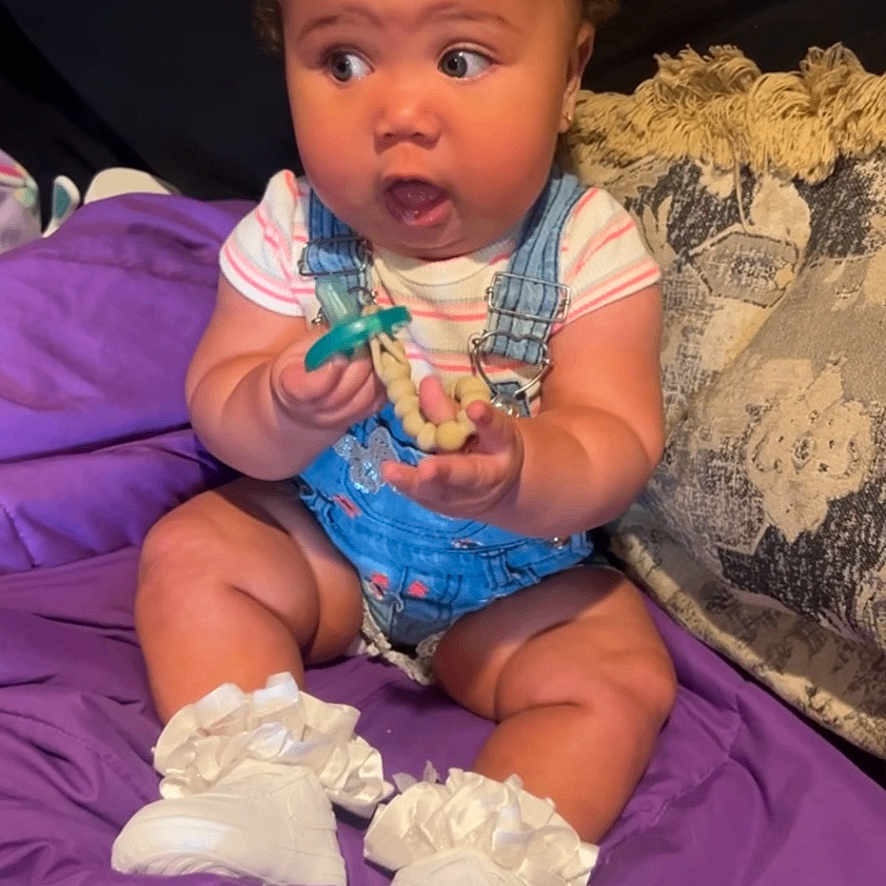 Alani joined the competition — help win amazing prizes! baby, child, infant, overalls, pacifier, beads, purple_blanket, couch, cushion, white_shoes, curly_hair, indoors, surprised_expression, sitting, clothing, person, cute, small_feet, home, portrait
