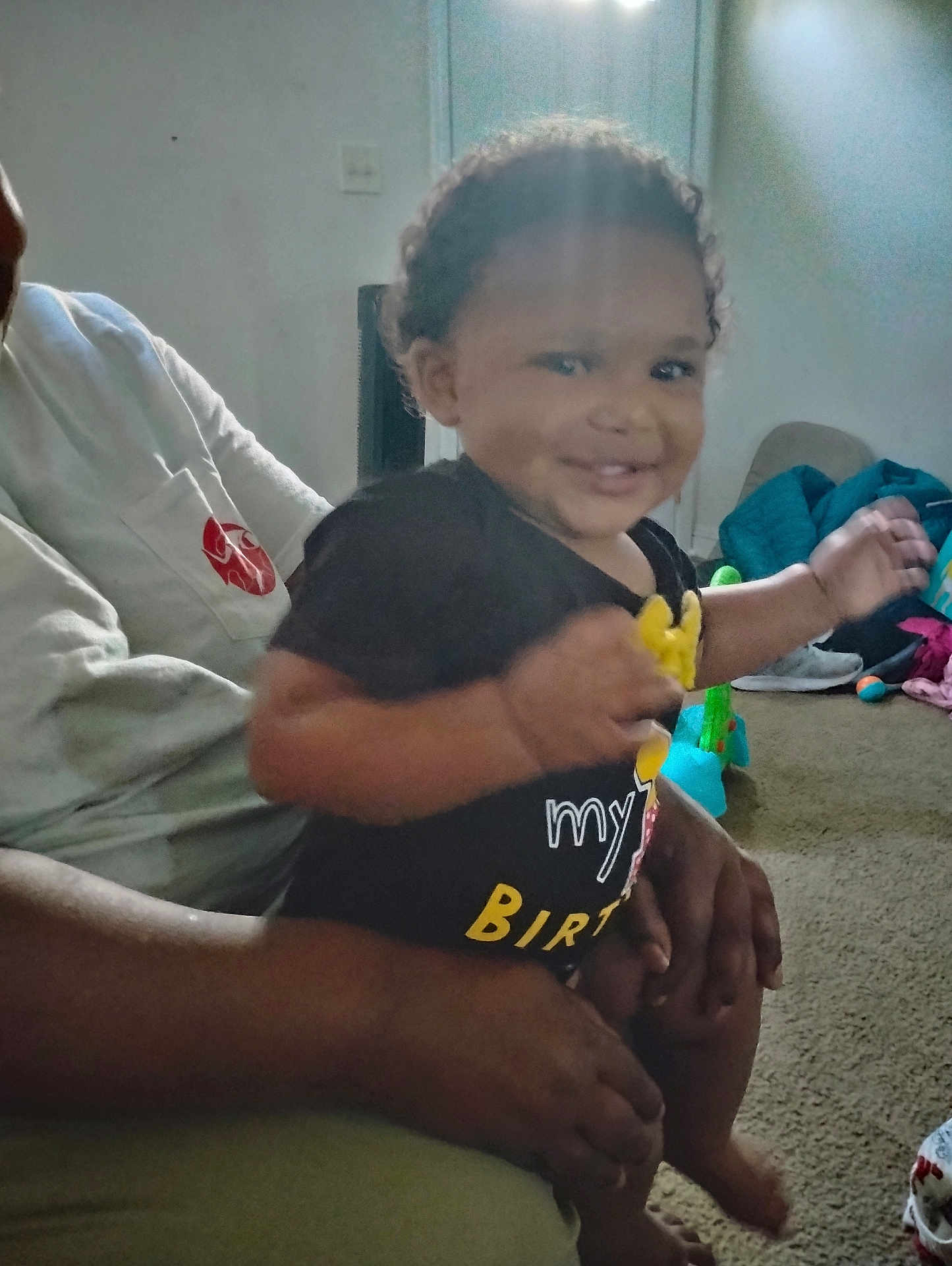 Eddonia Smith joined the competition — help win amazing prizes! toddler, child, smiling, person, adult, indoor, living_room, carpet, toy, clothing, black_shirt, hand, curly_hair, baby, happy, seated, family, playful, casual, home