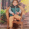 autumn, brown_shoes, casual_clothing, child, cute, fall_colors, fashion, forest_background, green_vest, indoor, photography, playful_pose, portrait, seated, smiling, studio_photo, tan_pants, toddler, wicker_chair, young_child