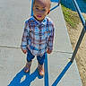 Bryston is registered to the contest to win money with this photo: bicycle, boots, boy, casual_clothing, child, concrete, curious, daylight, expression, outdoor, person, plaid_shirt, playground, railing, shadow, sidewalk, standing, sunny, urban, young