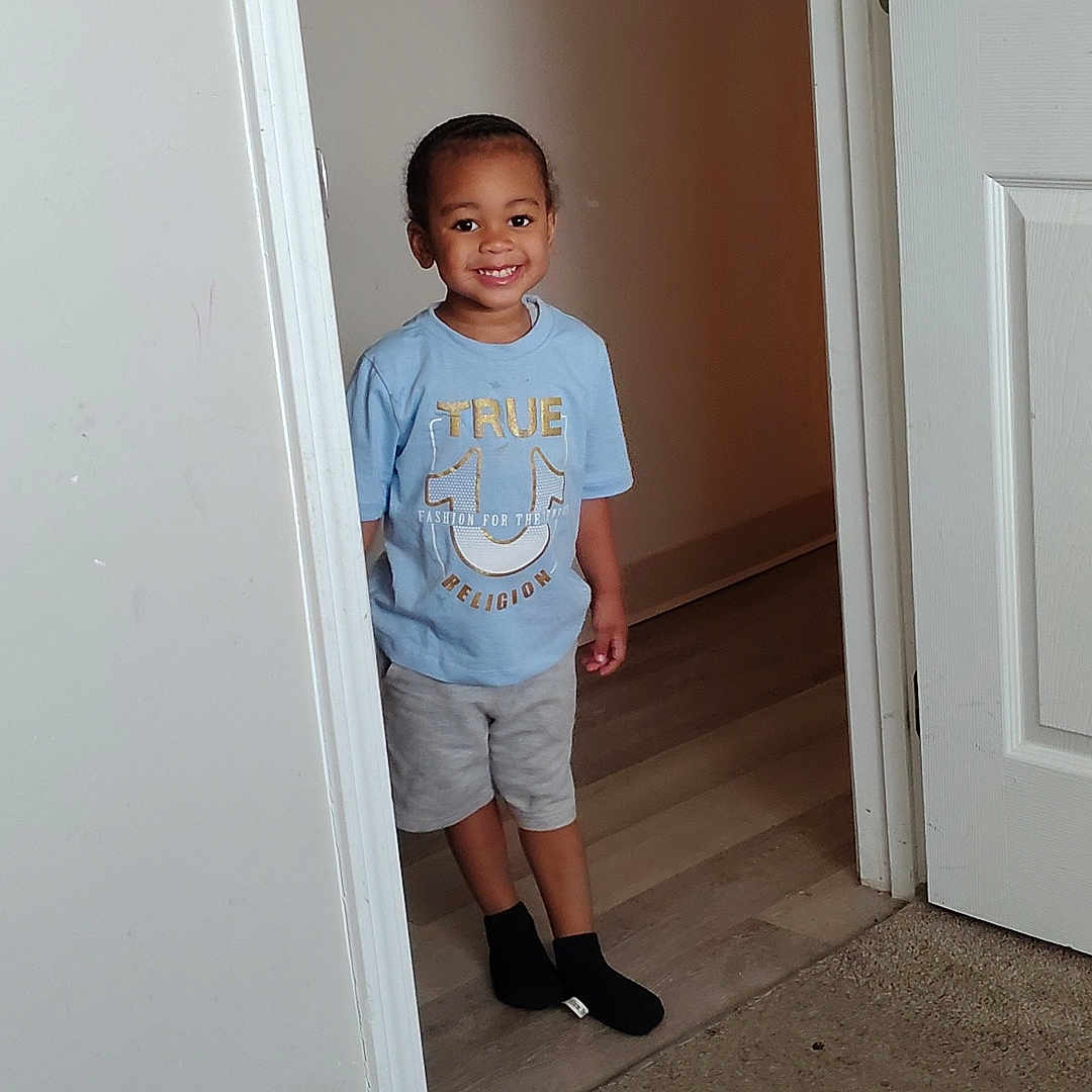 Bryston is registered to the contest to win money with this photo: blue_shirt, boy, carpet, casual_clothing, child, door, doorway, floor, happy, home, indoor, interior, person, portrait, shorts, smiling, socks, standing, wall, young_child