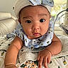 Adriana is registered to the contest to win money with this photo: baby, child, infant, portrait, floral_outfit, headband, bow, wide_eyed, indoors, couch, blanket, bedding, cozy, sitting, lying_down, face, hands, patterned_fabric, soft_lighting, home