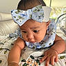 baby, infant, child, portrait, face, eyes, bow, headband, floral, patterned_clothing, bib, sitting, indoor, bed, blanket, furniture, hands, curious, close_up, home