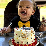 Mi'Zeir is registered to the contest to win money with this photo: birthday, cake, celebration, child, colorful, curly_hair, face, food, frosting, hands, happy, high_chair, messy, outdoor, party, person, smiling, table, toddler, yellow_bow_tie