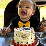 birthday, cake, celebration, child, colorful, curly_hair, face, food, frosting, hands, happy, high_chair, messy, outdoor, party, person, smiling, table, toddler, yellow_bow_tie